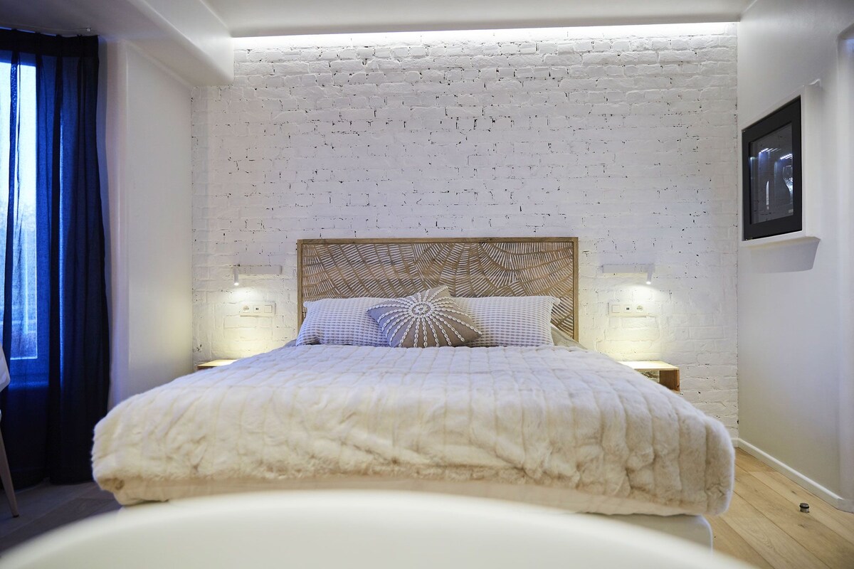 A bedroom with a white brick wall, a wooden headboard, two bedside lamps, and a bed with a white duvet.