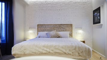 A bedroom with a white brick wall, a wooden headboard, two bedside lamps, and a bed with a white duvet.