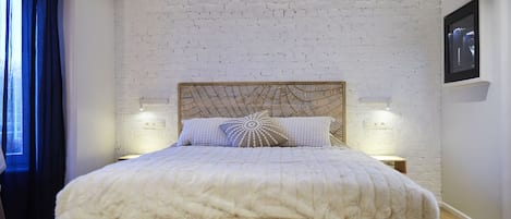 Quarto com parede de tijolos brancos, cabeceira de madeira, duas luminárias de cabeceira e cama com edredom branco.