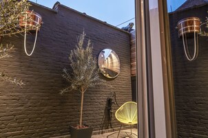 Un muro de ladrillo con un árbol en maceta, un espejo redondo y una silla amarilla.