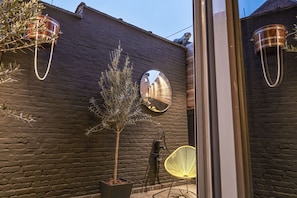 Un muro de ladrillo con un árbol en maceta, un espejo redondo y una silla amarilla.