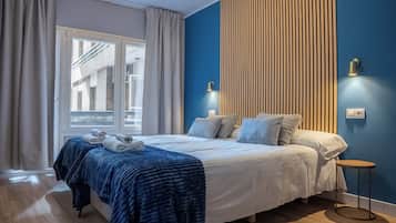 A modern bedroom with a large bed, a window with curtains, a blue accent wall, and a small side table.