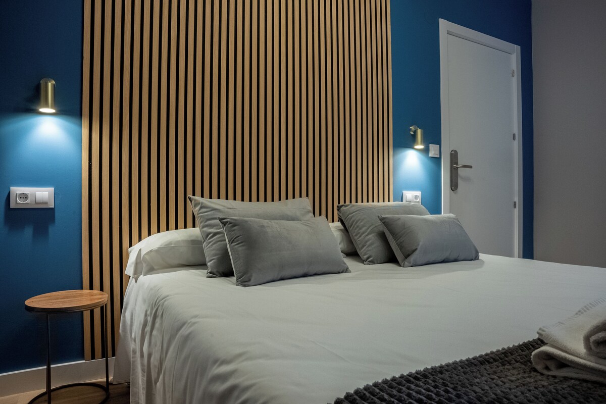 Een moderne slaapkamer met een bed, kussens, een nachtkastje en een wandlamp.
