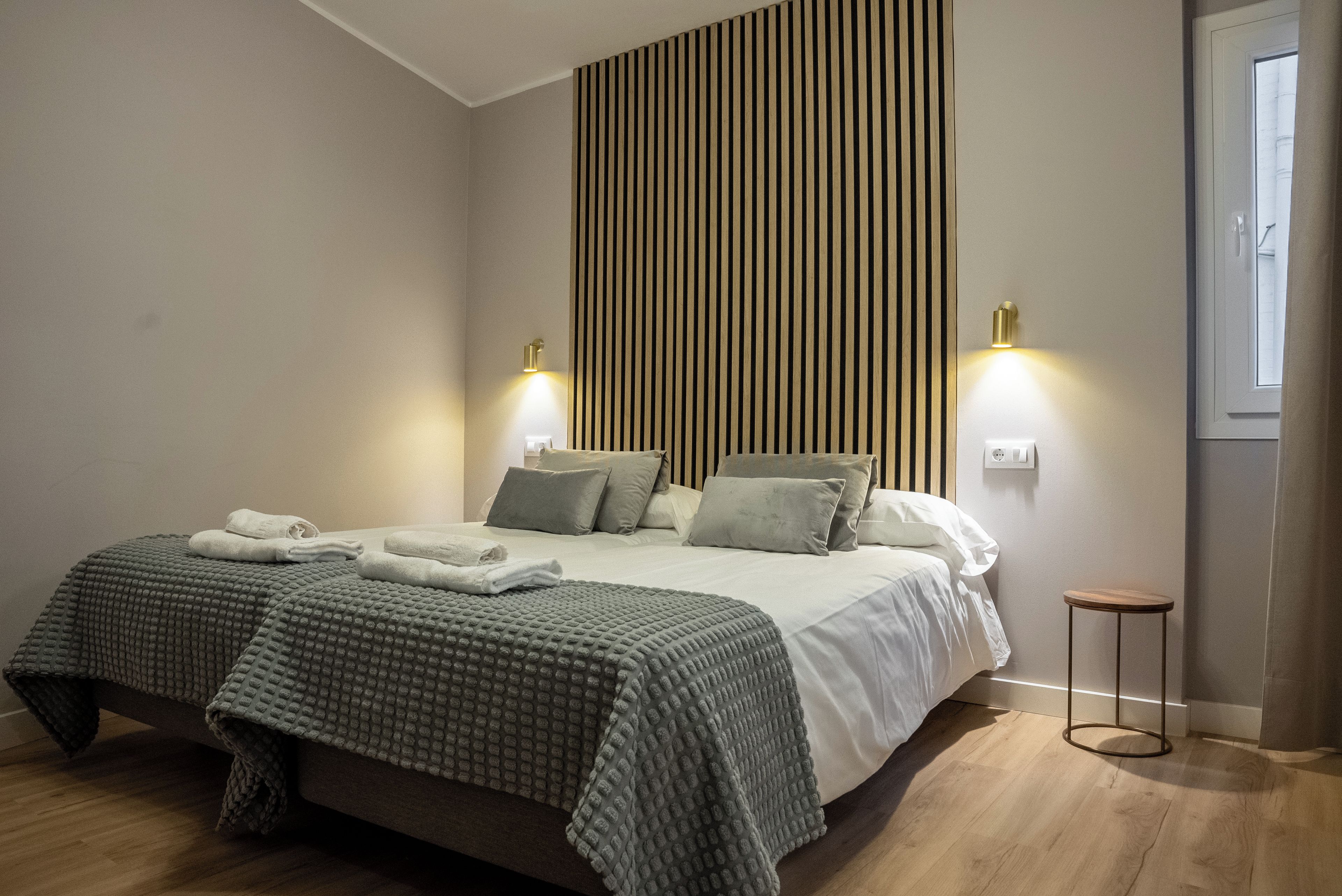 Quarto moderno com uma cama grande, duas luminárias de cabeceira, um banquinho pequeno e um painel de madeira vertical na parede.