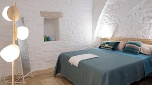 A bedroom with a stone wall, a bed with a blue bedspread, a bedside table with bottles, and a mirror with lights. - Ad Calcem "lagodentro" (Terlizzi)
