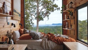 A cozy bedroom with a large window, wooden walls, a bed with pillows, a bedside table, and a shelf with decorative items. - Heyscape (Ocean Beach)