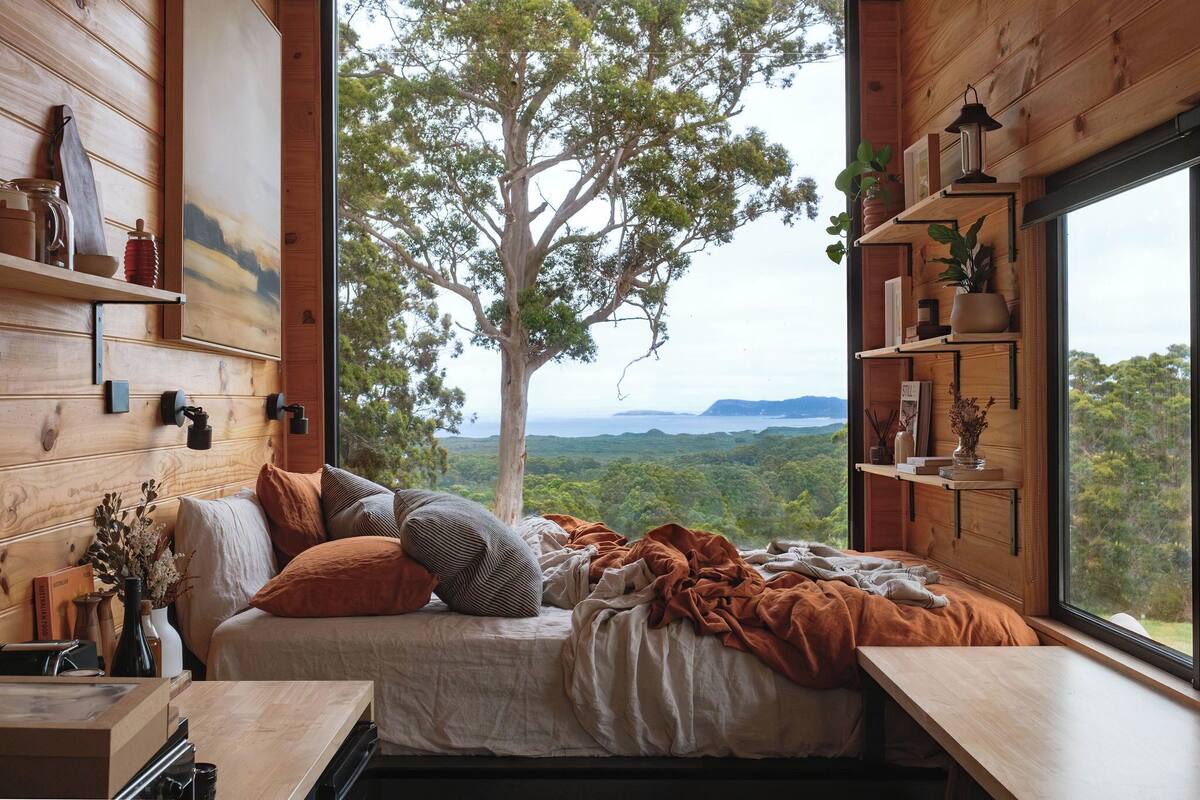 Une chambre confortable avec une grande fenêtre, des murs en bois, un lit avec des oreillers, une table de chevet et une étagère ornée d’objets décoratifs.