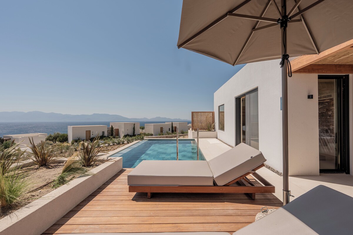A modern outdoor pool area with a wooden deck, lounge chairs, and a view of the sea.