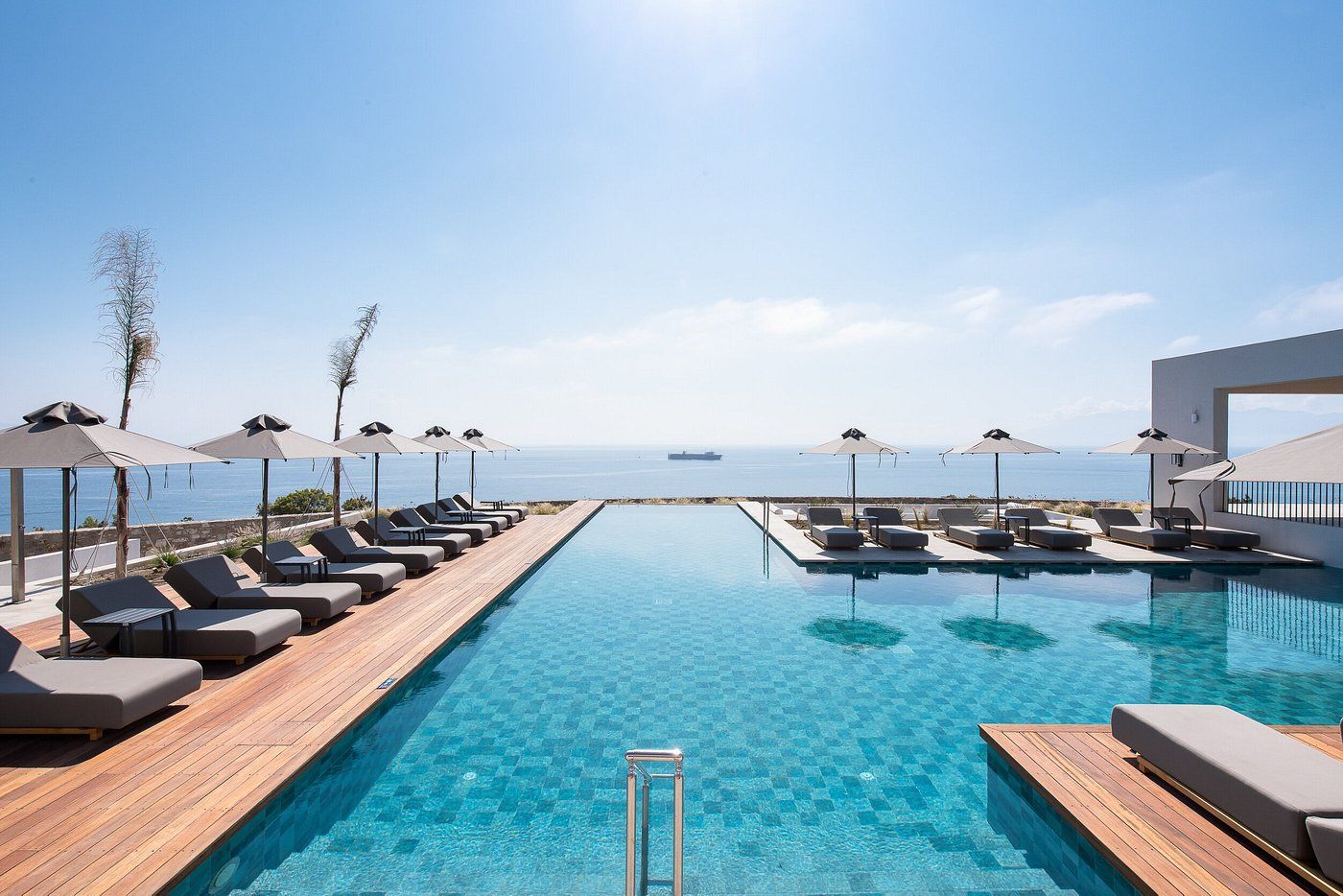 Un área junto a la piscina con sillones de descanso y sombrillas, una terraza de madera y vista al mar.