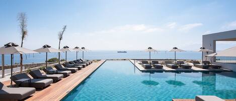 Ein Poolbereich mit Liegestühlen und Sonnenschirmen, ein Holzdeck und Blick aufs Meer.
