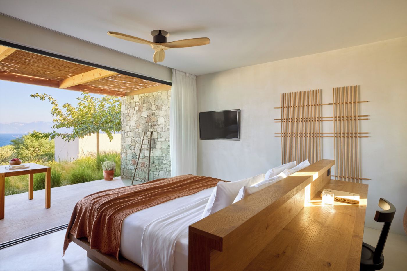 A modern bedroom with a large bed, wooden headboard, a ceiling fan, a stone wall, a TV, and a view of the outdoors.
