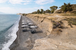 A beach with sun loungers and umbrellas, a rocky shoreline, and sparse vegetation. - Koia All-suite Wellbeing Resort (Kos)