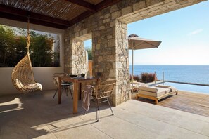 A stone patio with a hanging chair, a table with chairs, and a lounge area with a view of the sea. - Koia All-suite Wellbeing Resort (Kos)