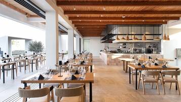 A spacious restaurant with long wooden tables and chairs, a kitchen area, and large windows allowing natural light.