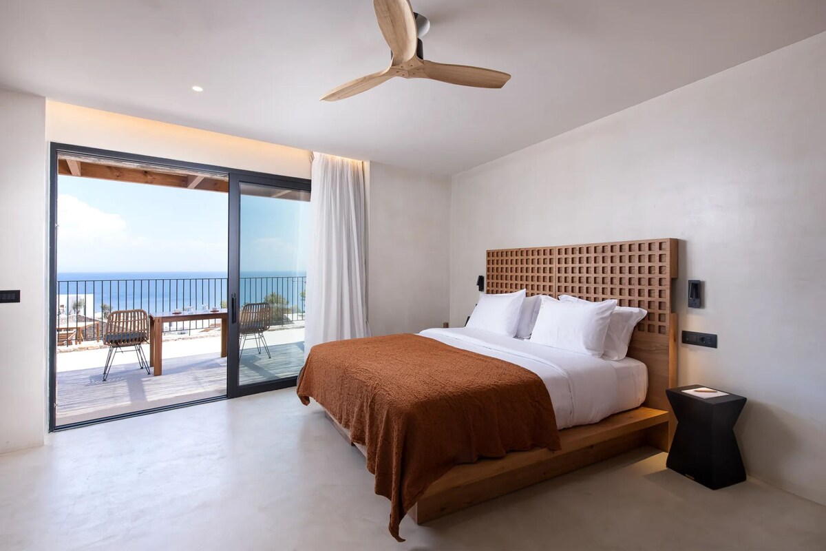 A bedroom with a bed, a ceiling fan, a view of the sea, and a balcony with outdoor furniture.