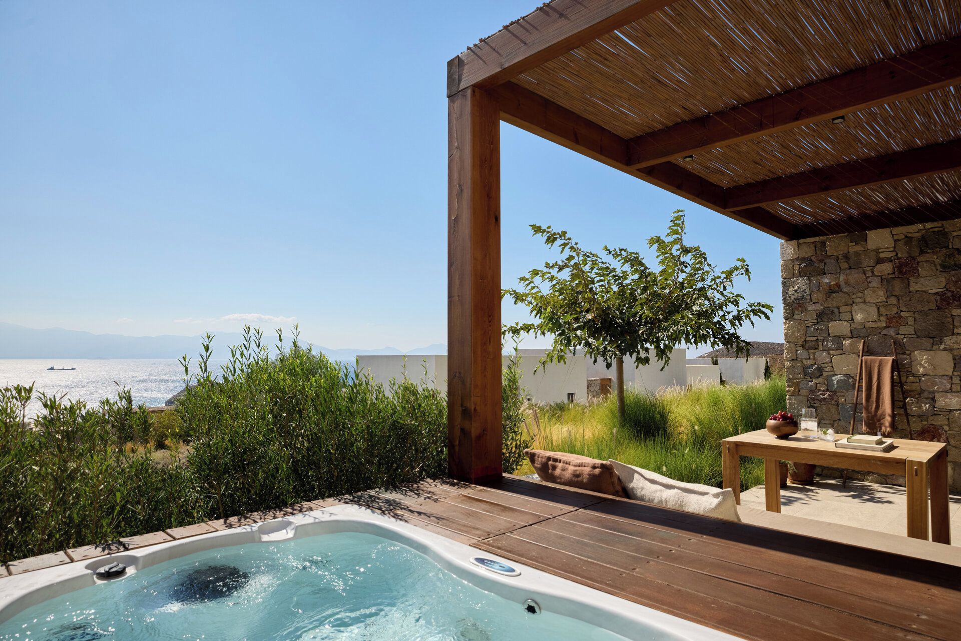 A hot tub with a wooden deck, a stone wall, and a view of the sea.