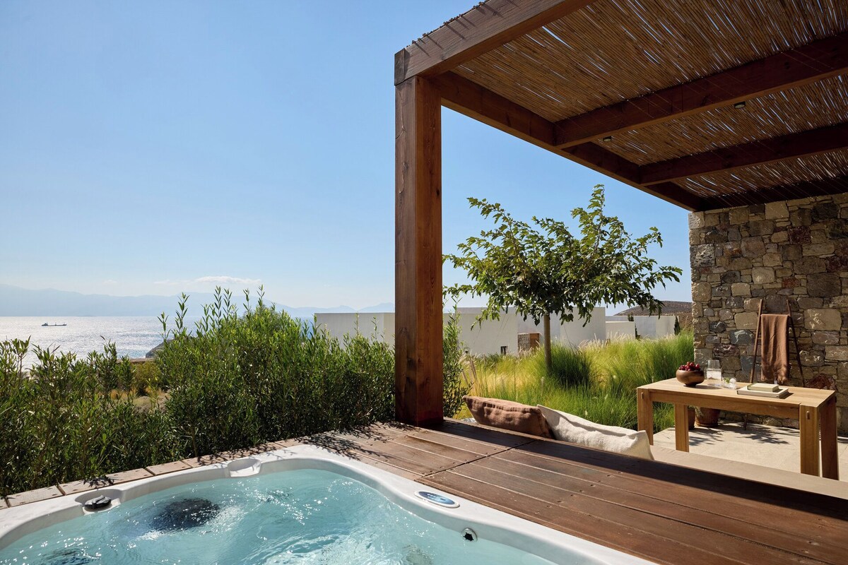 A hot tub with a wooden deck, a stone wall, and a view of the sea.