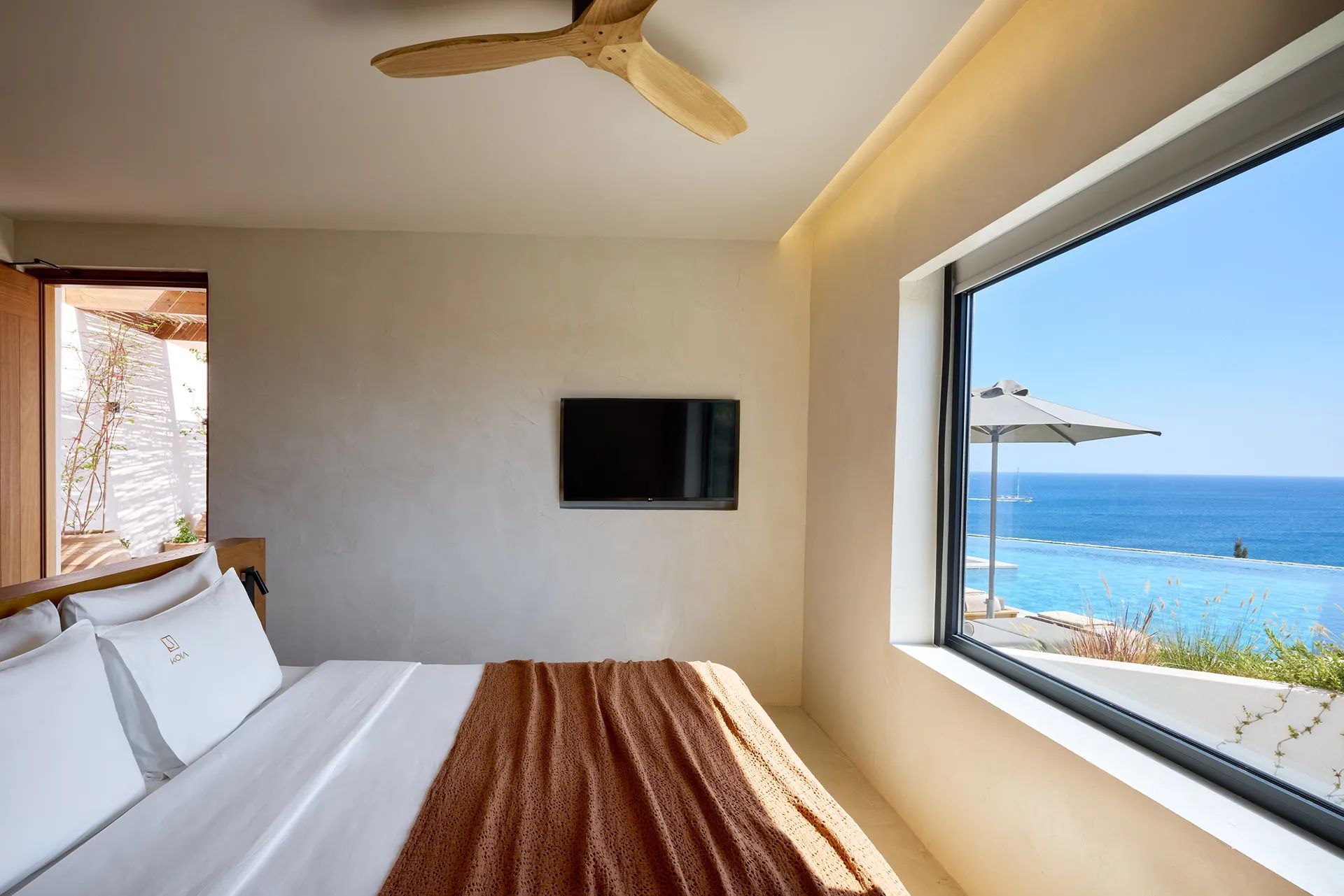 Quarto com cama, ventilador de teto, televisão e uma grande janela com vista para o oceano.