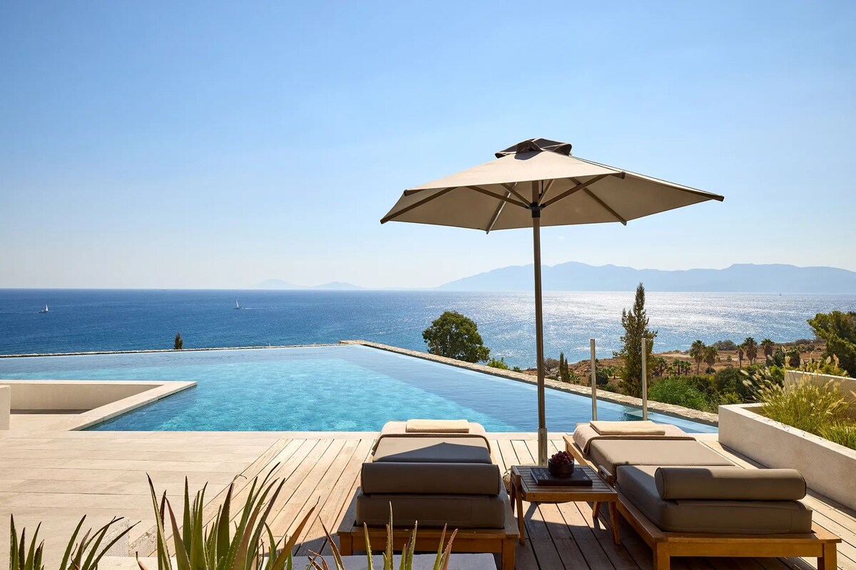 A poolside area with lounge chairs and an umbrella, overlooking a body of water with a distant mountain range.