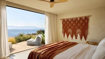 A bedroom with a large bed, a ceiling fan, a macrame wall hanging, and a view of the sea.