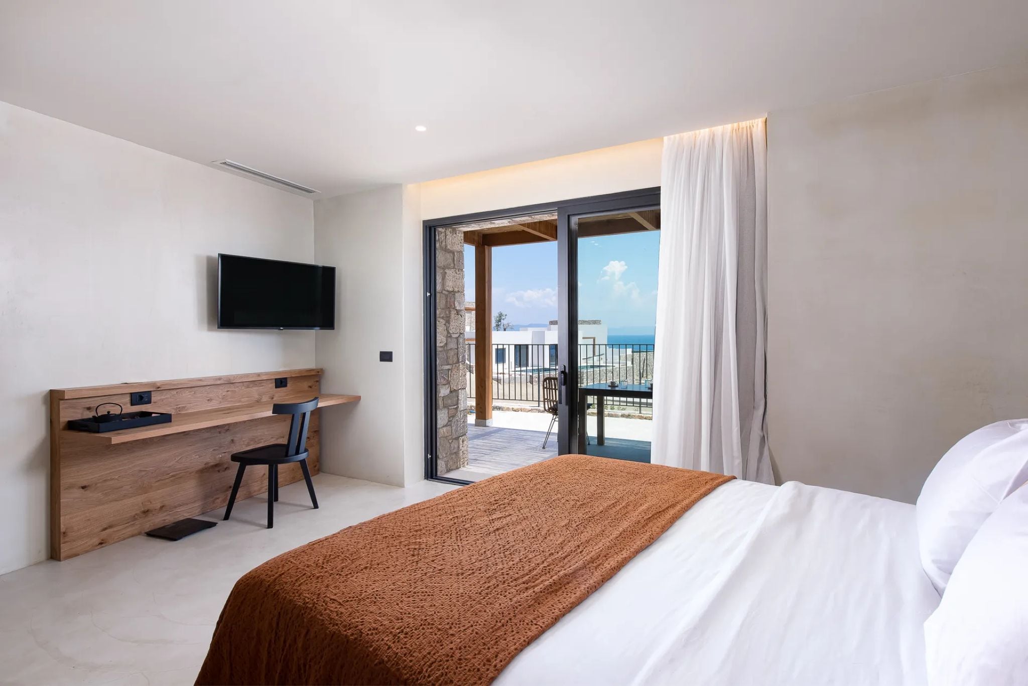 A hotel room with a bed, a desk, a chair, a television, and a balcony with a view.