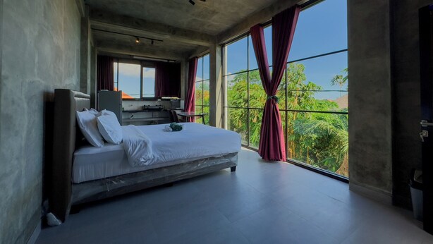 A modern bedroom with a large bed, a desk, and a view of greenery through large windows. - Sky Apartments And Suites By Zuzu (Pererenan)