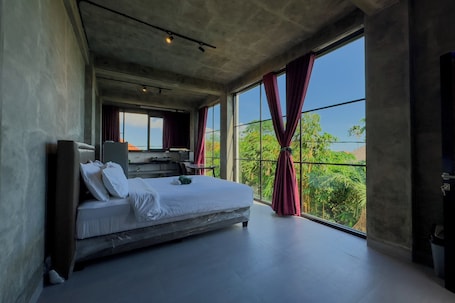 A modern bedroom with a large bed, a desk, and a view of greenery through large windows.