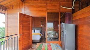 A wooden cabin with a balcony, a bed with a white headboard, a dining area with a table and chairs, and a refrigerator. - Sky Apartments And Suites By Zuzu (Pererenan)