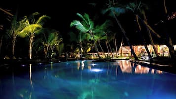 Un espace piscine illuminé la nuit, agrémenté de palmiers et où les lumières se reflètent sur l’eau.