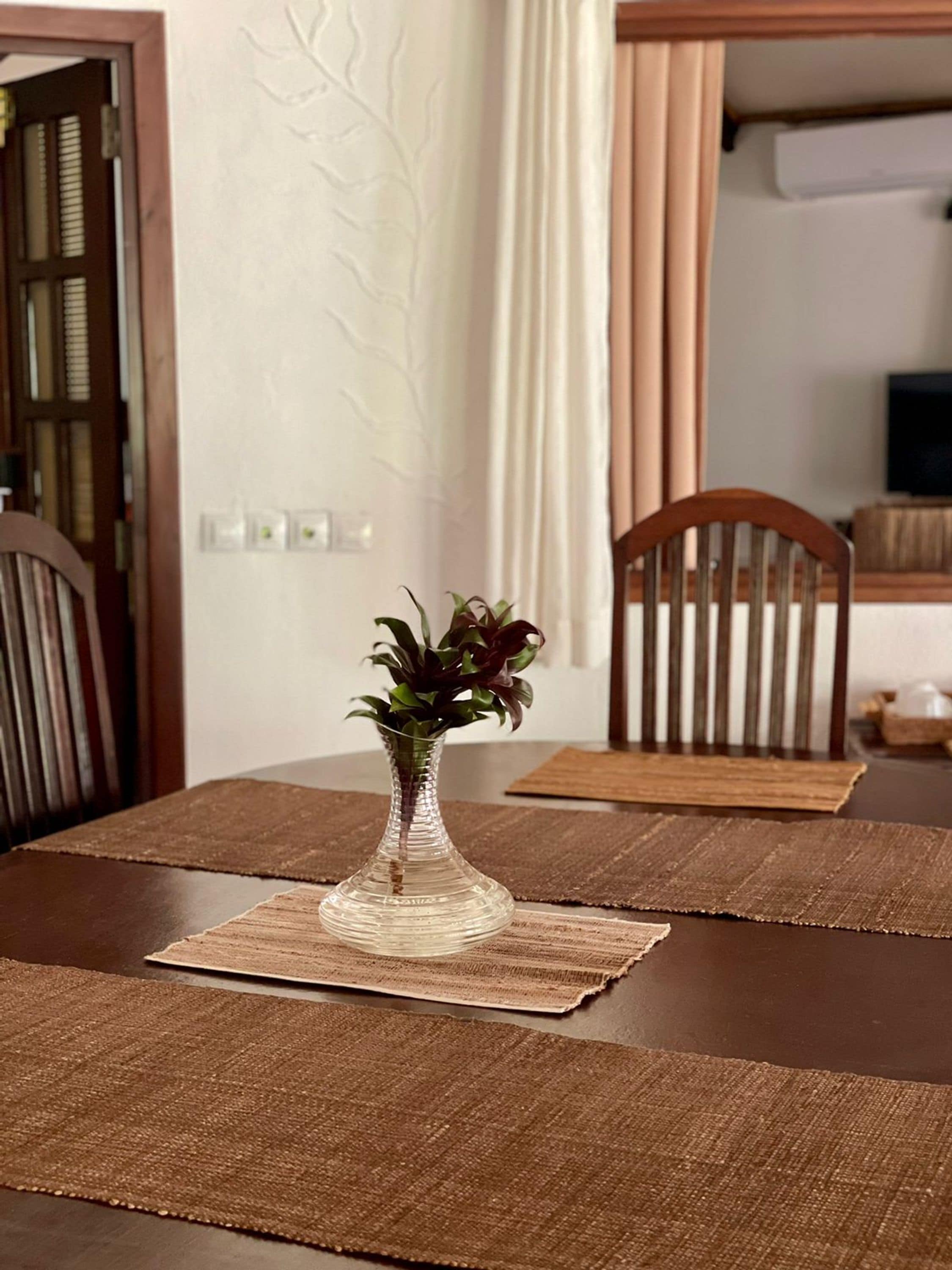 A dining table with a clear glass vase containing a plant, set on a brown placemat.