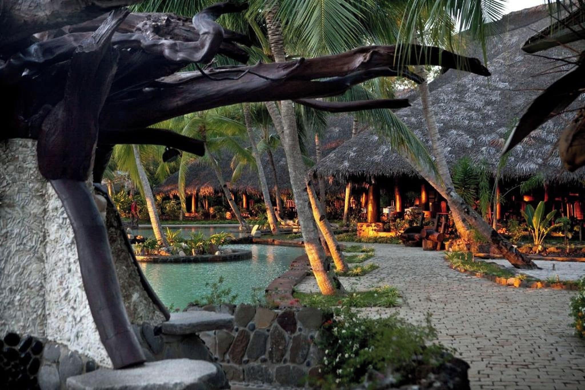 Een tropisch resort met een zwembad, palmbomen en overkappingen met rieten daken.