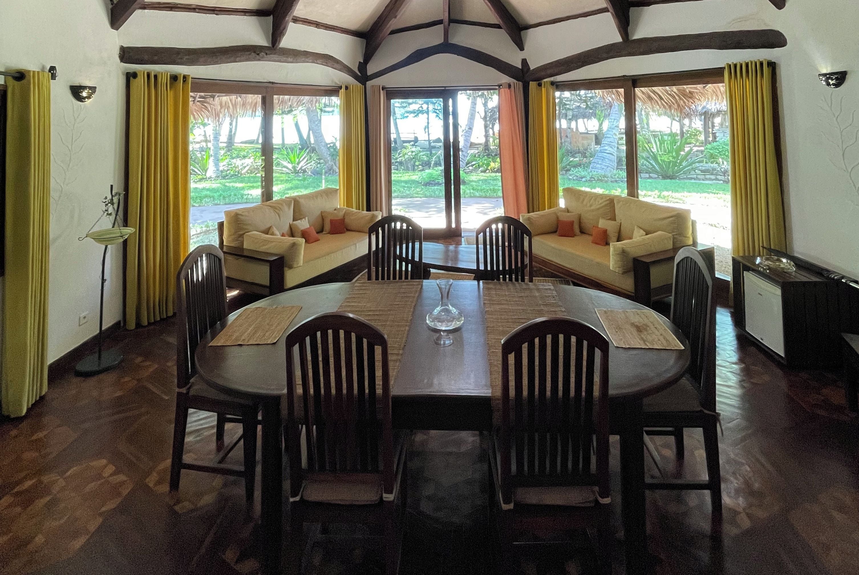 Un comedor con una mesa de madera, sillas y un sofá. Hay cortinas en las ventanas y se ve una lámpara.