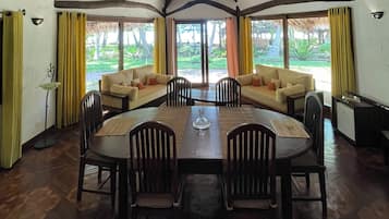 Un comedor con una mesa de madera, sillas y un sofá. Hay cortinas en las ventanas y se ve una lámpara.