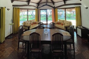 A dining area with a wooden table, chairs, and a sofa. There are curtains on the windows, and a lamp is visible. - Ravintsara Wellness Hotel (Nosy Be)