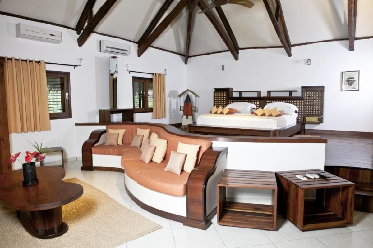 Una habitación acogedora con un sofá curvo, una pequeña mesa de madera, una cama con cabecero de madera y una ventana con cortinas.
