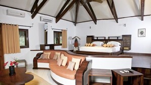 A cozy room with a curved sofa, a small wooden table, a bed with a wooden headboard, and a window with curtains. - Ravintsara Wellness Hotel (Nosy Be)