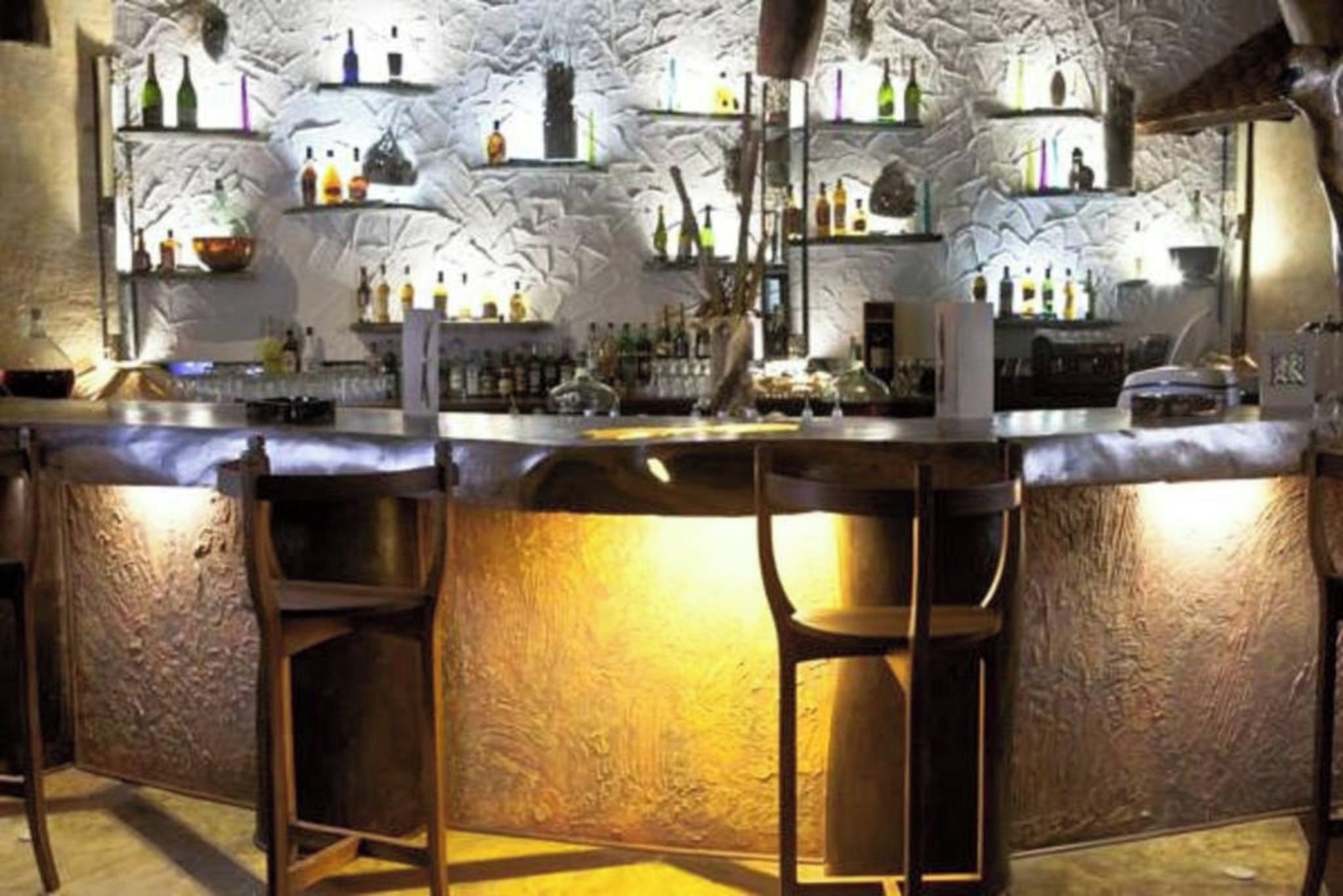 A bar with a rustic stone wall, shelves with bottles, and bar stools.