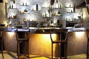 Un bar con pared de piedra rústica, estantes con botellas y taburetes de bar.