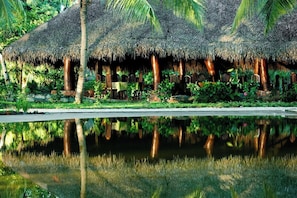 A thatched-roof structure with chairs and tables, surrounded by lush greenery and a reflective water body. - Ravintsara Wellness Hotel (Nosy Be)