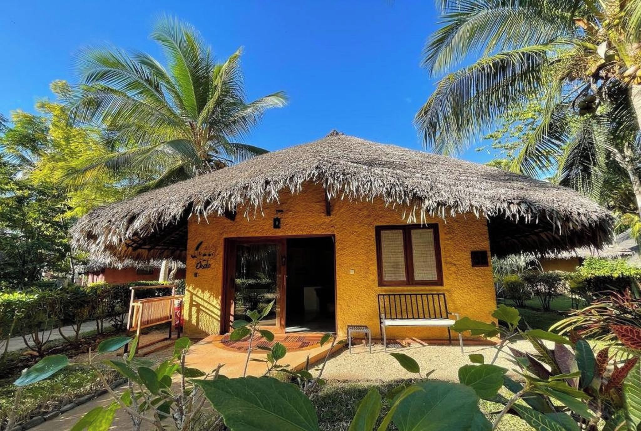 A small, single-story building with a thatched roof, surrounded by lush greenery and palm trees.