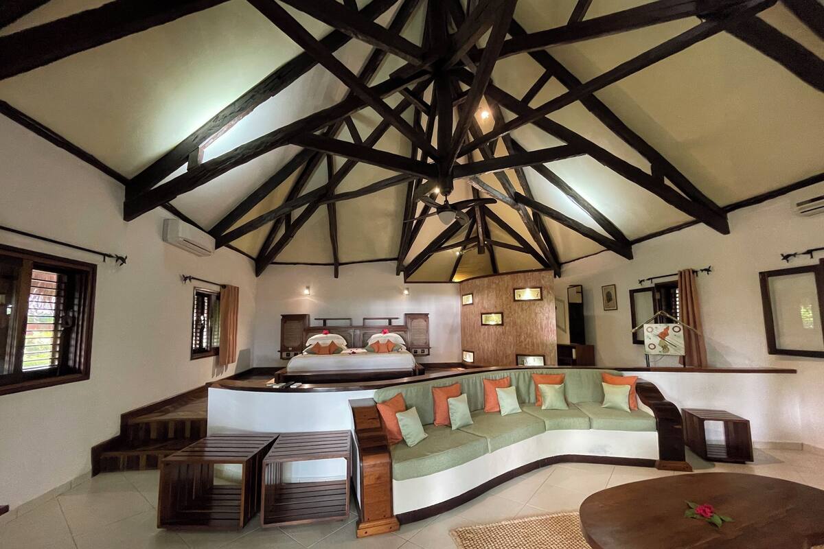 A spacious room with a high ceiling, a curved green sofa, a wooden coffee table, and a bed with a wooden headboard.