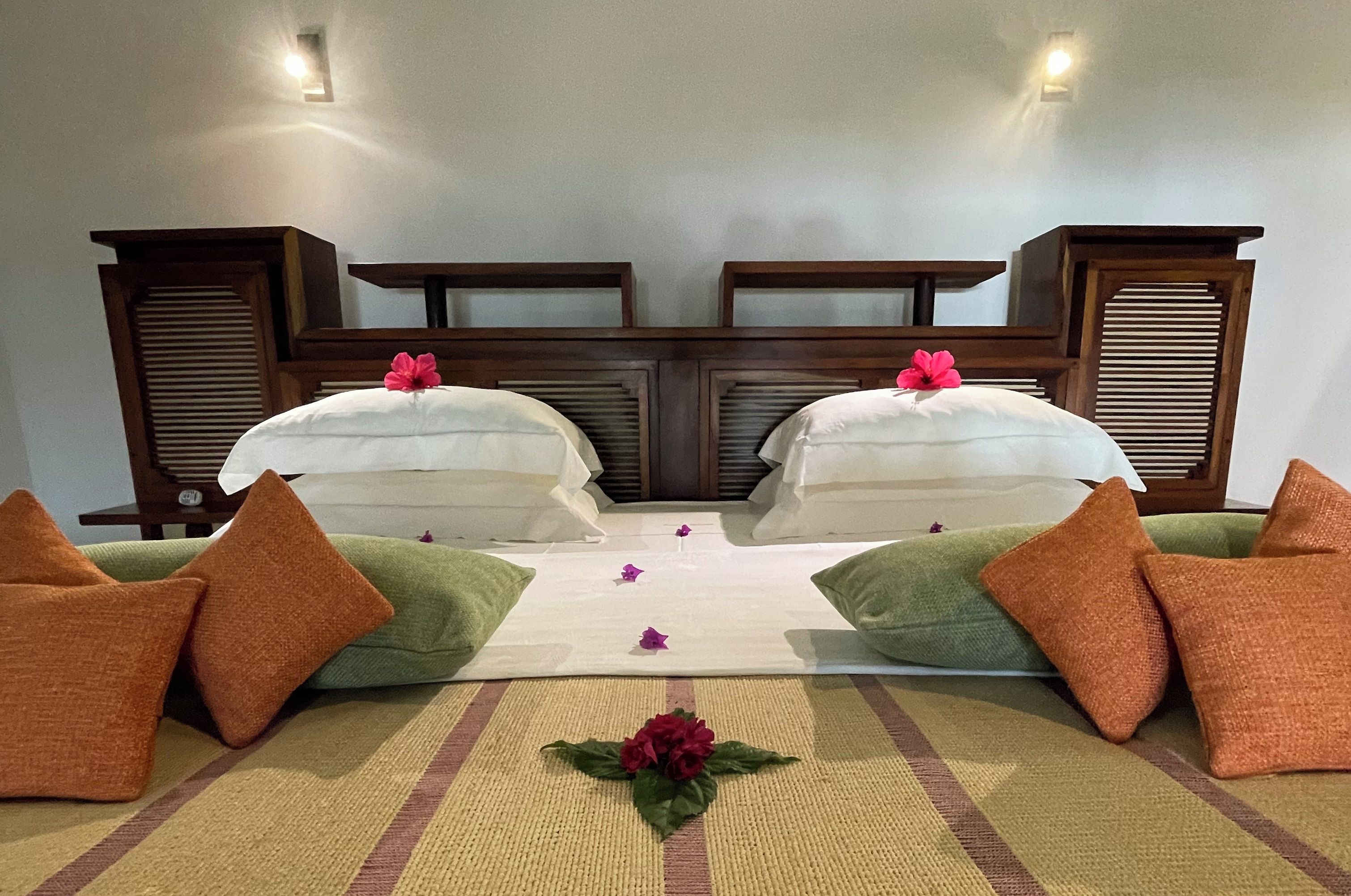 Un letto matrimoniale con lenzuola bianche, due testiere in legno, cuscini verdi e arancioni e un composizione floreale decorativa sul letto.