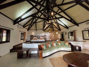 A spacious room with a high ceiling, a curved green sofa, a wooden coffee table, and a bed with a wooden headboard. - Ravintsara Wellness Hotel (Nosy Be)