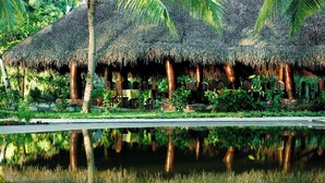 A thatched-roof structure with chairs and tables, surrounded by lush greenery and a reflective water body. - Ravintsara Wellness Hotel (Nosy Be)
