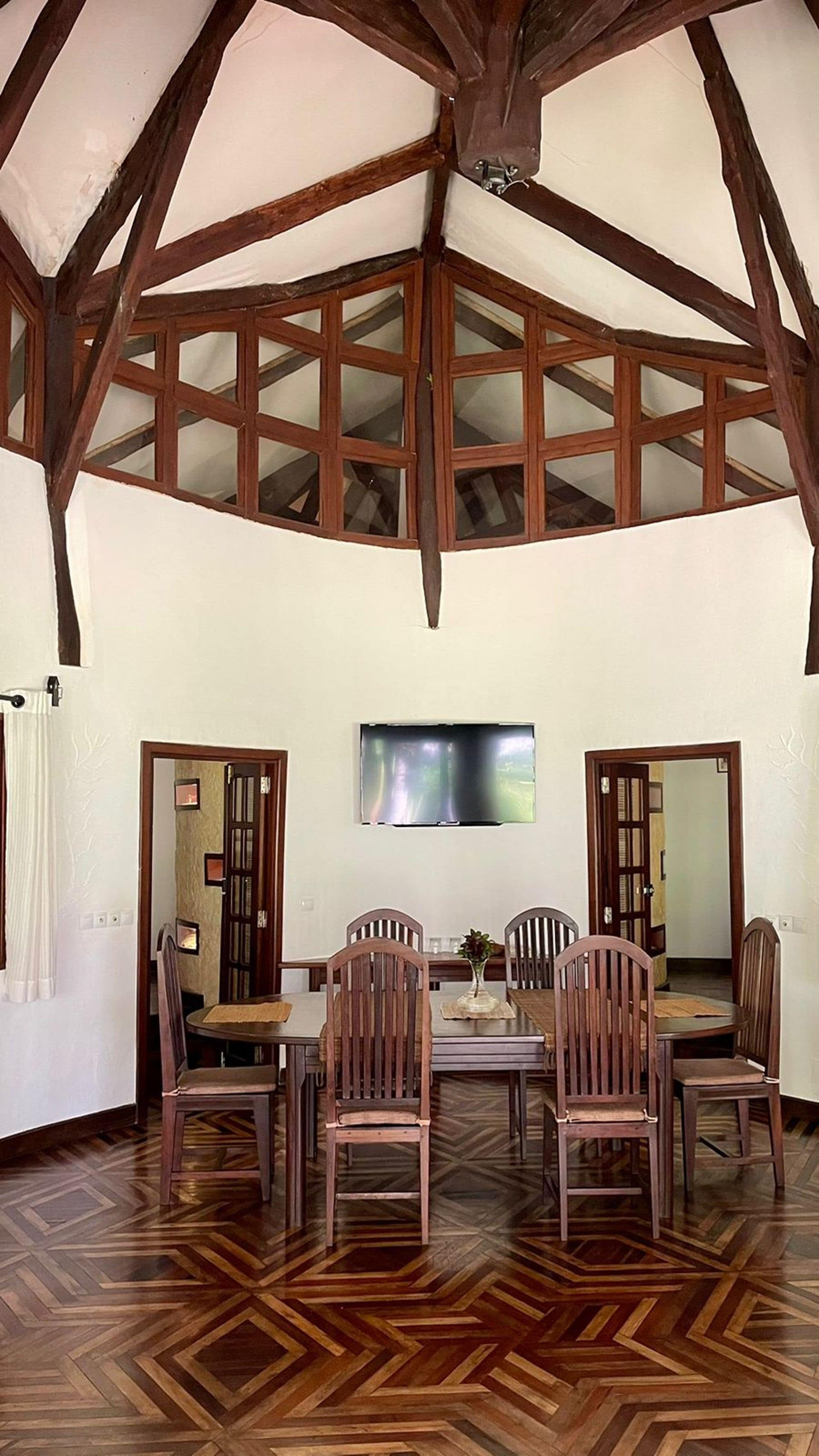 Un espace repas doté d’un plafond haut et de poutres apparentes, une table centrale entourée de chaises et un téléviseur fixé au mur.