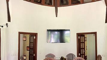 A dining area with a high ceiling and exposed wooden beams, a central table with chairs, and a television mounted on the wall.