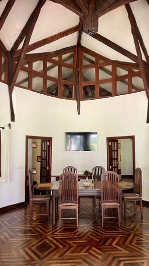 A dining area with a high ceiling and exposed wooden beams, a central table with chairs, and a television mounted on the wall. - Ravintsara Wellness Hotel (Nosy Be)