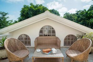 A patio area with wicker furniture, a table with a teapot and a book, and a white building with arched windows in the background. - Elivaas Monforte Ayada - Luxury Villa In Goa (Assagao)