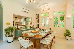 A dining area with a wooden table set for six, surrounded by chairs. There is a kitchen area with a counter and a mirror above it. The room has a ceiling fan and a window with green foliage visible through the glass. - Elivaas Monforte Ayada - Luxury Villa In Goa (Assagao)