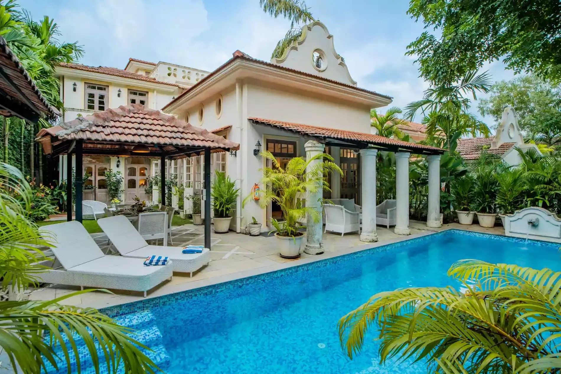 A pool area with lounge chairs, a covered seating area, and a building with a white facade and red-tiled roof.